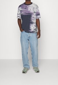 Muž v tričku s abstraktním vzorem v purpurové a bílé barvě, v modrých volných džínách a zelených teniskách, stojící s jednou rukou v kapse.