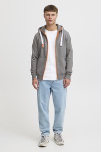 Sudadera con cremallera gris con detalles en naranja, con capucha ajustable y bolsillos delanteros. Lleva puesta sobre una camiseta blanca y combinada con vaqueros azul claro.