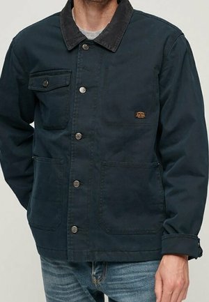 Mann trägt eine dunkelblaue Jacke mit Knöpfen, einem schwarzen Kragen, drei Vordertaschen und hellblaue Jeans.