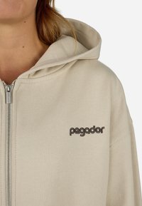 Corta-vento de cor creme com fecho de correr, feito de tecido suave, apresentando um capô com cordão e o logótipo "pegador" bordado em castanho no peito.