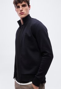 Νέος άνδρας φορώντας μαύρο μπουφάν με φερμουάρ και ψηλό γιακά πάνω από μαύρο πουκάμισο και μπεζ παντελόνι, σε απλό λευκό φόντο.