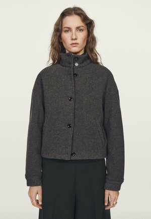 Veste cropped gris foncé avec un col haut, dotée de boutons noirs et de poignets côtelés. Tissu doux et texturé avec une coupe décontractée.
