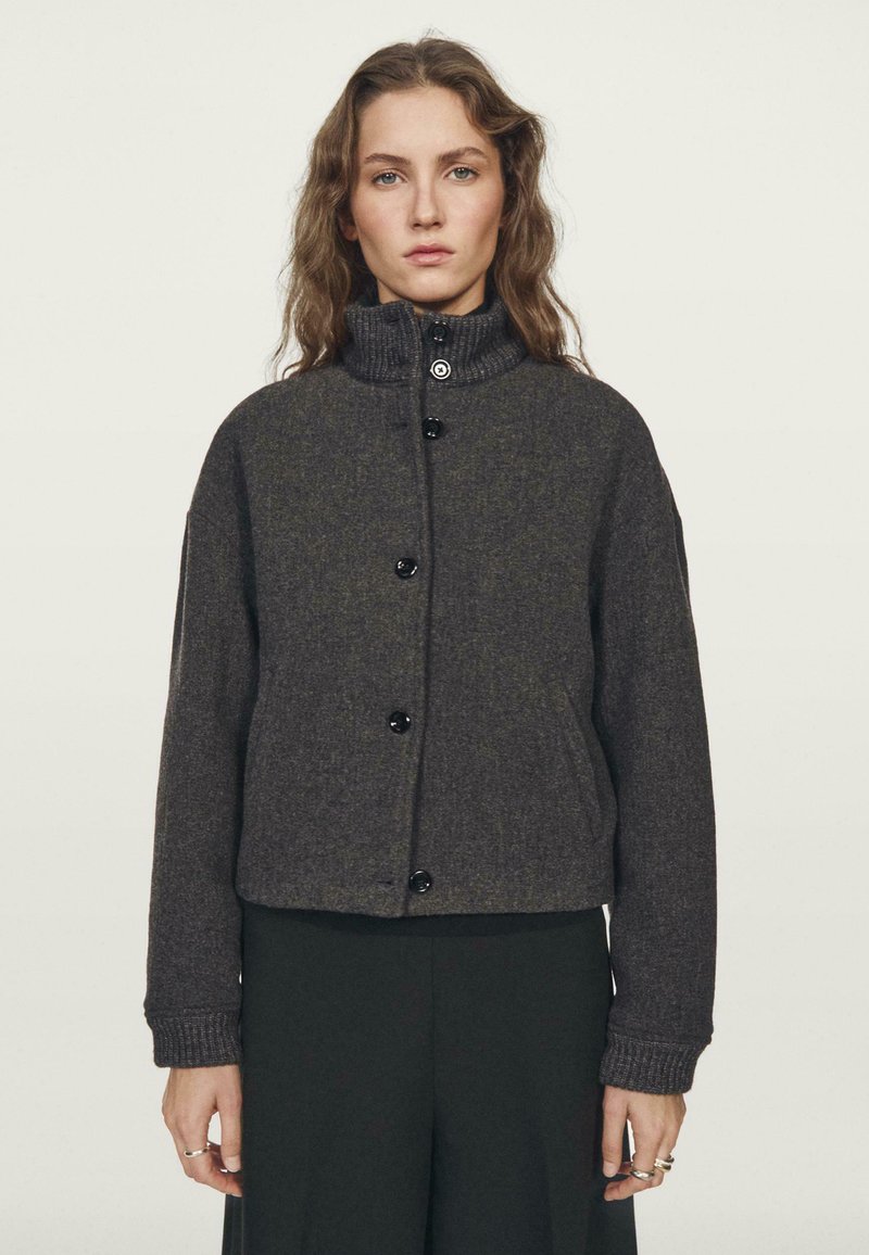 Veste cropped gris foncé avec un col haut, dotée de boutons noirs et de poignets côtelés. Tissu doux et texturé avec une coupe décontractée.