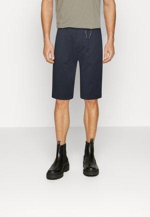 Homme portant un short bleu marine au genou, des bottines en cuir noires et une chemise grise, debout devant un fond blanc uni.