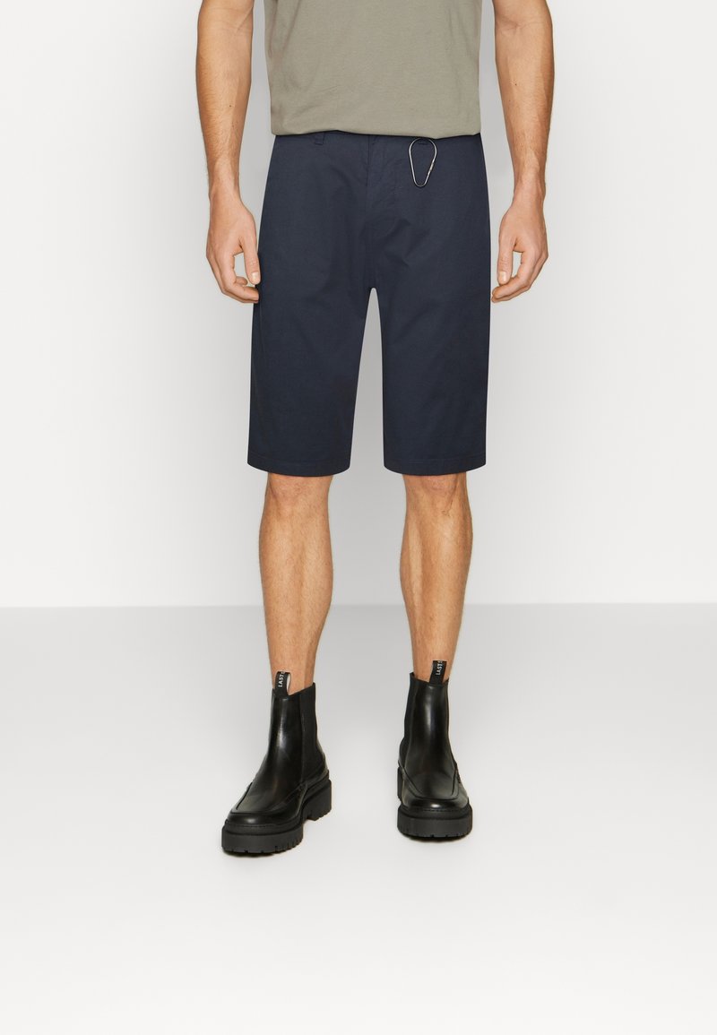 Homme portant un short bleu marine au genou, des bottines en cuir noires et une chemise grise, debout devant un fond blanc uni.