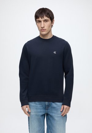 Ung mand iført en navy sweatshirt med lille hvidt "cK" logo på brystet og lyseblå jeans, stående mod en ensfarvet hvid baggrund.