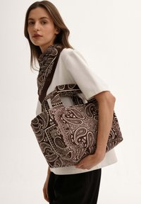 Kobieta z długimi brązowymi włosami nosi białą bluzkę z krótkim rękawem, brązowy szal w wzór paisley oraz niesie dużą torbę w pasującym wzorze paisley.