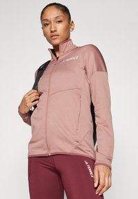 Svijetlo ružičasta zip-up jakna s visokim ovratnikom i bočnim džepovima, izrađena od teksturirane tkanine; nosi se s odgovarajućim bordo tajicama i crnim ruksakom.