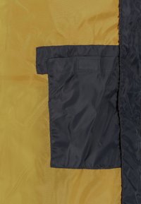Žlutá látka s hladkým povrchem má námořnickou kapsu. Kapsa má šitý okraj a uzávěr na suchý zip.