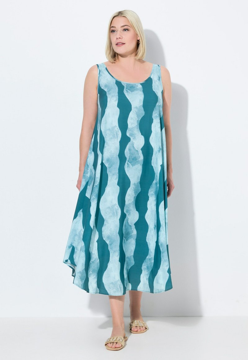 Ärmelloses türkisfarbenes Kleid mit wellenförmigem, hellblauem Muster, fließender Silhouette, in der Mitte der Wadenlänge, aus weichem Stoff, kombiniert mit hellen Sandalen.