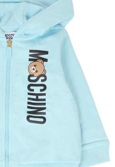 Jasnoniebieska kurtka z kapturem, zapinana na zamek, wykonana z miękkiego materiału. Posiada graficzny nadruk z kreskówkowym niedźwiedziem oraz wyraźny czarny napis "Moschino". Rękawy i dół zakończone ściągaczem.