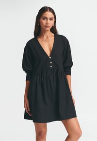Schwarzes Baumwollkleid mit tiefem V-Ausschnitt, dreiviertel langen Puffärmeln und gerafftem Taillenbund, ausgestattet mit drei dekorativen Knöpfen.