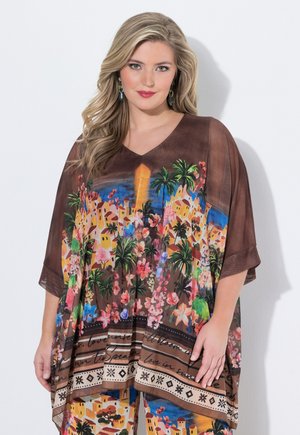 Vaaleahiuksinen nainen pukeutuneena ruskeaan, löysään toppiin ja housuihin, joissa on värikkäitä kukka- ja kylämalleja, seisoo valkoisen taustan edessä.