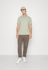 Lichtgroen poloshirt met korte mouwen, gecombineerd met een bruine broek en witte sneakers. De man draagt een lichtbeige pet.