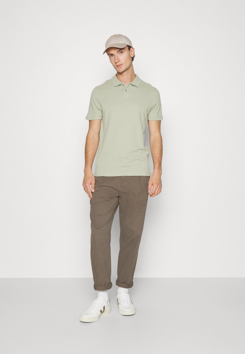 Lichtgroen poloshirt met korte mouwen, gecombineerd met een bruine broek en witte sneakers. De man draagt een lichtbeige pet.