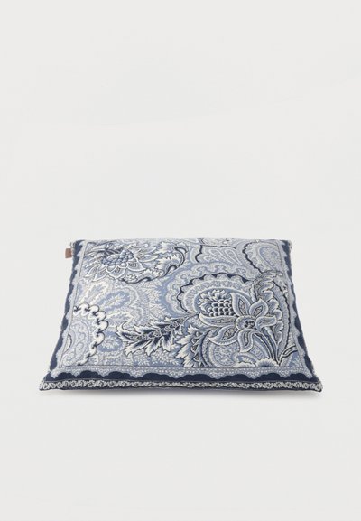 Coussin carré avec un motif complexe de fleurs bleues et blanches en forme de paisley et une bordure festonnée bleu foncé sur un fond uni.