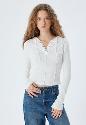 Top blanco de manga larga con capucha, que presenta una abertura con botones y detalles cosidos. Combinado con jeans de mezclilla azul.