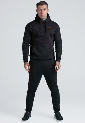 Mann som står med hendene ved siden av seg, iført svart hettegenser med gull emblem, svarte joggebukser og svarte joggesko på lys grå bakgrunn.