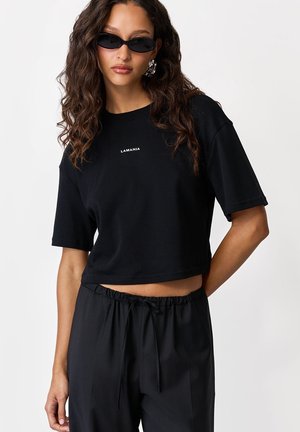 Kobieta z długimi, kręconymi włosami, nosząca czarny krótki t-shirt z napisem "LAMANIA", czarne spodnie ze sznurkiem, czarne okulary przeciwsłoneczne i srebrne kolczyki.