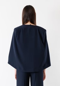 Grembiule di colore blu navy, in stile mantella, con una vestibilità rilassata, rifinito con un collo arrotondato e un piccolo dettaglio con bottone sul retro. Tessuto dalla consistenza liscia.