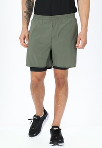Pantaloni sportivi verde oliva con vita elastica, tessuto liscio e un pantaloncino interno nero a strati, indossati con scarpe da corsa nere.
