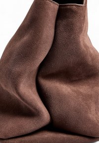 Brun mocka handväska med en mjuk, texturerad yta och en avslappnad design. Den har en bred öppning och minimalistiska metallaccenter.