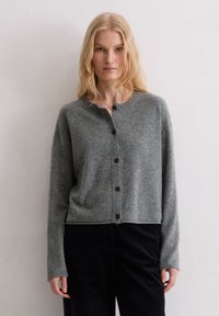 Cardigan en tricot gris avec un col rond, une fermeture à boutons, des poignets côtelés et une coupe courte, porté sur un pantalon en velours côtelé noir.
