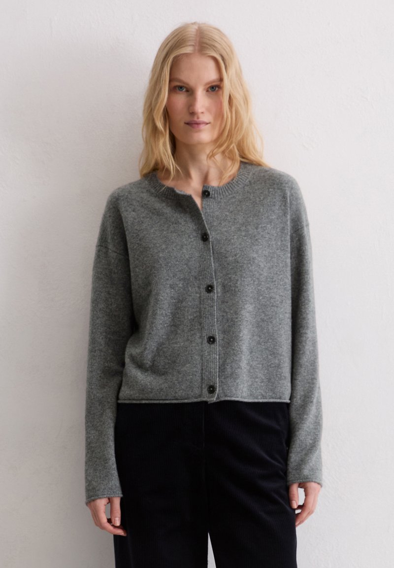 Cardigan en tricot gris avec un col rond, une fermeture à boutons, des poignets côtelés et une coupe courte, porté sur un pantalon en velours côtelé noir.