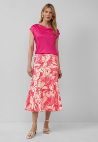 Haut drapé fuchsia à manches courtes, associé à une longue jupe rose ornée d'un motif floral dans des teintes de rouge et de rose, accentué par des sandales blanches.