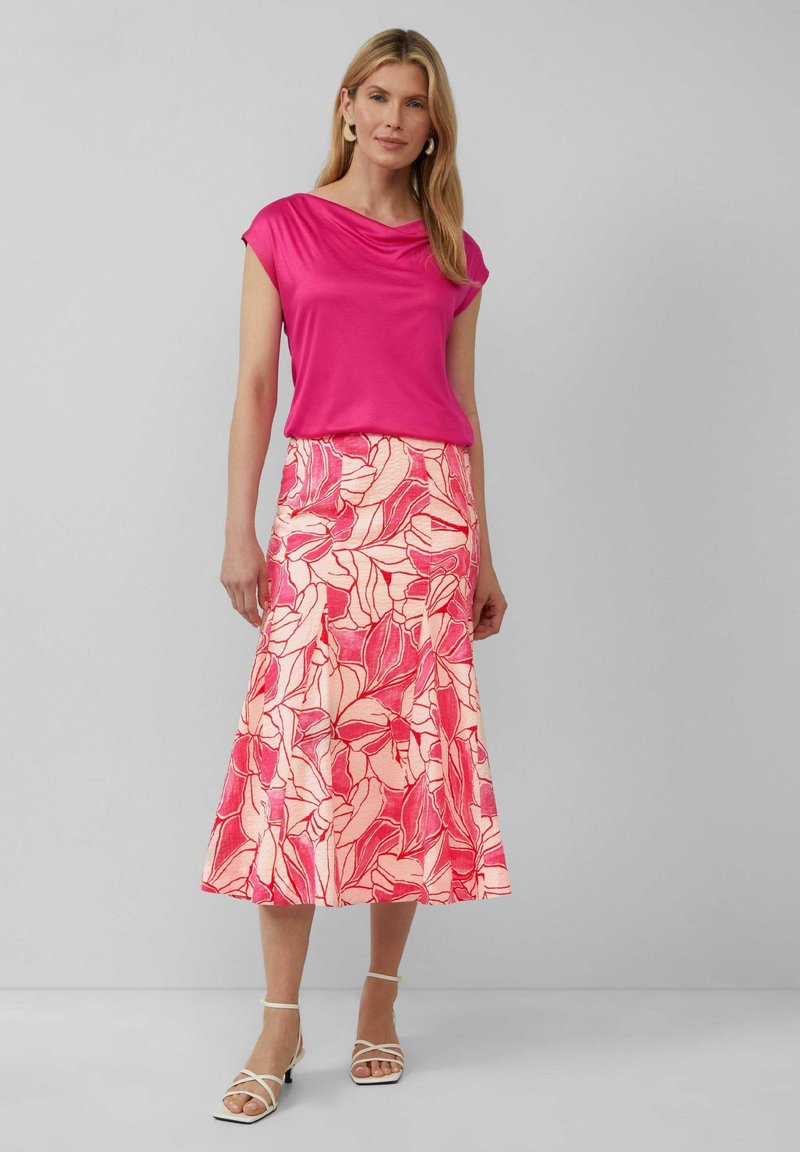 Haut drapé fuchsia à manches courtes, associé à une longue jupe rose ornée d'un motif floral dans des teintes de rouge et de rose, accentué par des sandales blanches.