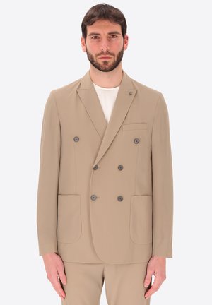 Uomo con capelli corti e scuri e barba che indossa una giacca beige doppiopetto sopra una camicia bianca su sfondo bianco.