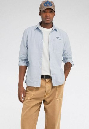 Junger Mann mit kariertem Cap, hellblau gestreiftem Hemd über weißem T-Shirt und beiger Hose, stehend vor einfarbigem Hintergrund.