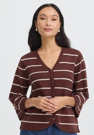 Vrouw met lang donker haar, met gouden oorringen en een bruine cardigan met knopen en witte horizontale strepen, staand tegen een effen achtergrond.