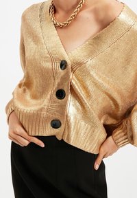 Goldener, metallischer Cardigan mit V-Ausschnitt, gerippten Bündchen und schwarzen Knöpfen. Glatte Textur mit dezentem Schimmer, kombiniert mit schwarzer Hose.