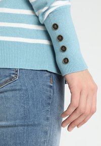 Jasnoniebieski sweter w paski o ribbowym wykończeniu, z czterema czarnymi guzikami na mankiecie i dopasowanym kroju, zestawiony z niebieskimi dżinsami.