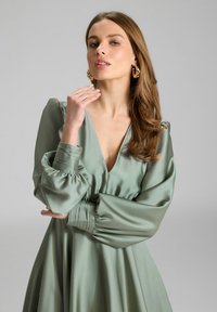 Robe en satin vert avec un décolleté en V profond, des manches bouffantes et des poignets plissés. Texture lisse et design fluide, rehaussé par de grandes boucles d'oreilles en or.