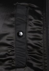 Tissu noir avec un rabat rectangulaire cousu, maintenu par un seul bouton noir sur une surface lisse et brillante.
