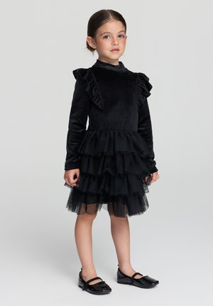 Abito nero con maniche a volant, gonna in tulle a strati e corpetto in velluto. Completato da eleganti ballerine nere.