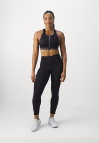 Schwarzer Sport-BH mit einem Reißverschluss vorne und einem Logo-Band, kombiniert mit hochgeschnittenen schwarzen Leggings. Das Model trägt weißeSportschuhe.