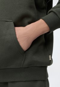Main placée dans la poche avant d'un sweat-shirt vert foncé à manches et ourlet côtelés, montrant une partie du pantalon assorti.