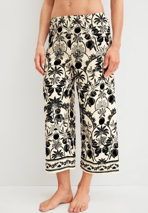 Beige broek met wijde pijpen, bedrukt met zwarte bloemen- en fruitpatronen, gedragen door een persoon die op blote voeten staat met ontspannen handen langs het lichaam.