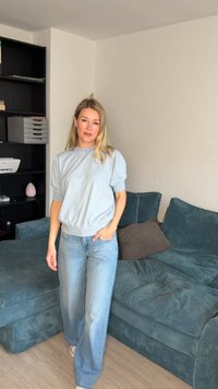 Γυναίκα με ανοιχτό μπλε πουλόβερ και τζιν, στέκεται στο σαλόνι κοντά σε τυρκουάζ καναπέ με μαξιλάρια και μαύρη μονάδα ραφιών.