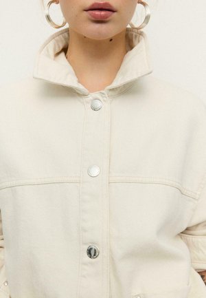 Žena v svetlobéžovej košeľovej bunde s gombíkmi, so zvýšeným golierom a veľkými kruhovými náušnicami, vidieť čiastočne tvár a hornú časť trupu.