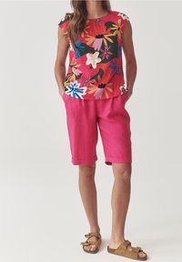 Oberteil mit floralem Muster in lebendigen Farben, kombiniert mit pinken Shorts. Das Outfit zeichnet sich durch eine entspannte Passform und leichtes Material aus, ergänzt durch braune Sandalen.