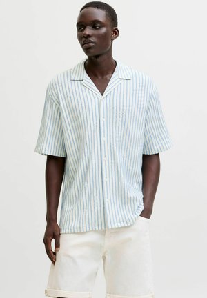 Jeune homme portant une chemise blanche à manches courtes avec des rayures verticales bleues et un short blanc arrivant aux genoux, debout devant un fond uni.