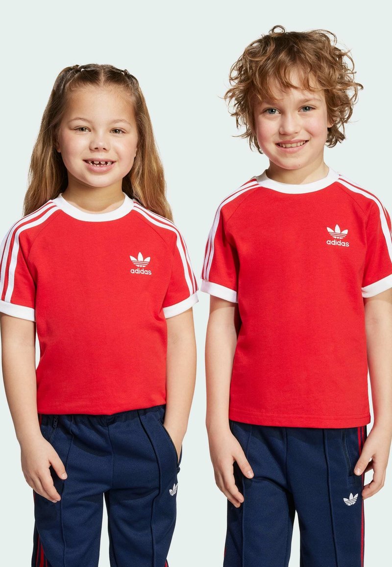 Red cotton t-shirts with white trim and Adidas logo, worn by two children. One shirt has a round neckline; both feature striped shoulders.