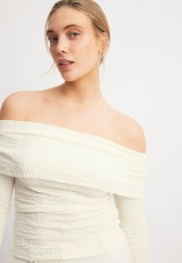Off-Shoulder-Oberteil aus strukturiertem, elfenbeinfarbigem Stoff mit einem zerknitterten Design und langen Ärmeln, das mit gerafften Details für eine taillierte Passform versehen ist.