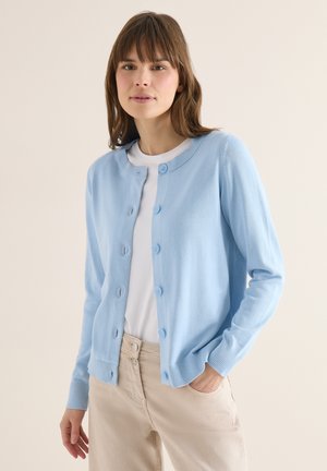 Jeune femme portant un cardigan bleu clair boutonné sur une chemise blanche et un pantalon beige, debout avec une main dans la poche sur un fond uni.