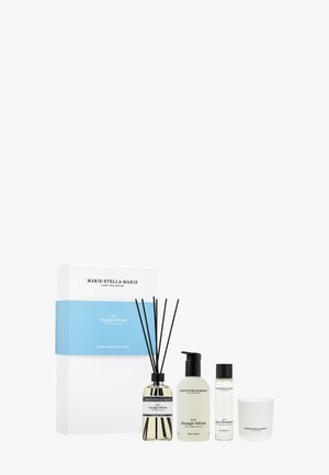 Ensemble de parfum d'intérieur comprenant un diffuseur en verre avec des tiges noires, un gel douche, un gel nettoyant et une bougie, le tout dans un emballage blanc et transparent.