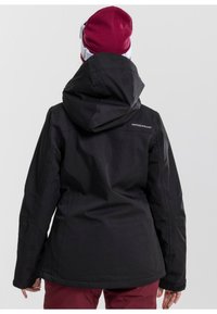 Schwarze Outdoor-Jacke mit Kapuze, mit einer taillierten Silhouette und verstellbaren Manschetten. Logo auf dem oberen Rücken. Kombiniert mit bordeauxroten Hosen.